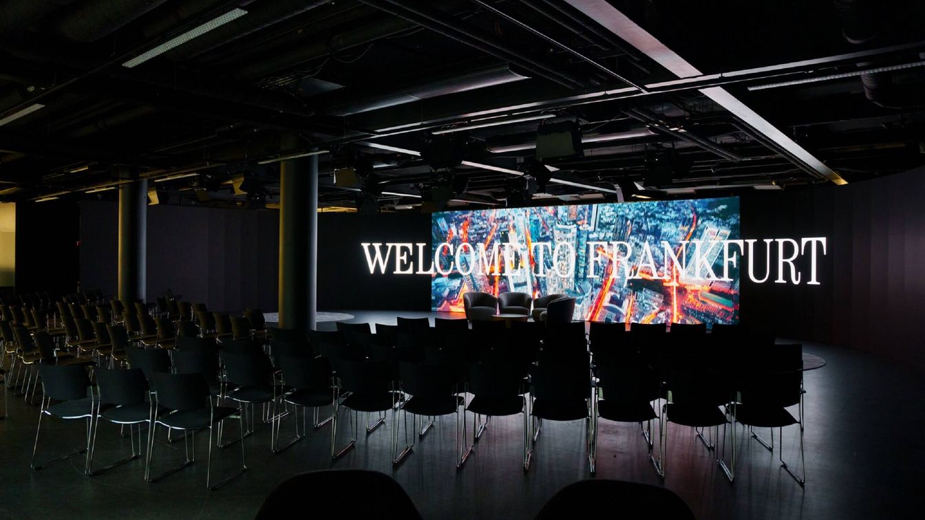 Stuhlreihen stehen vor einem großen Screen mit der Aufschrift Welcome in Frankfurt.